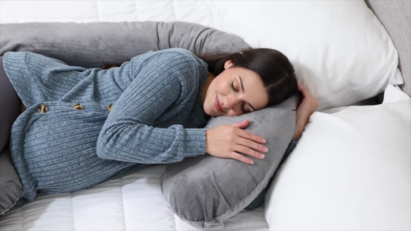 Trudnica leži na boku, zagrljena sa velikim sivim jastukom